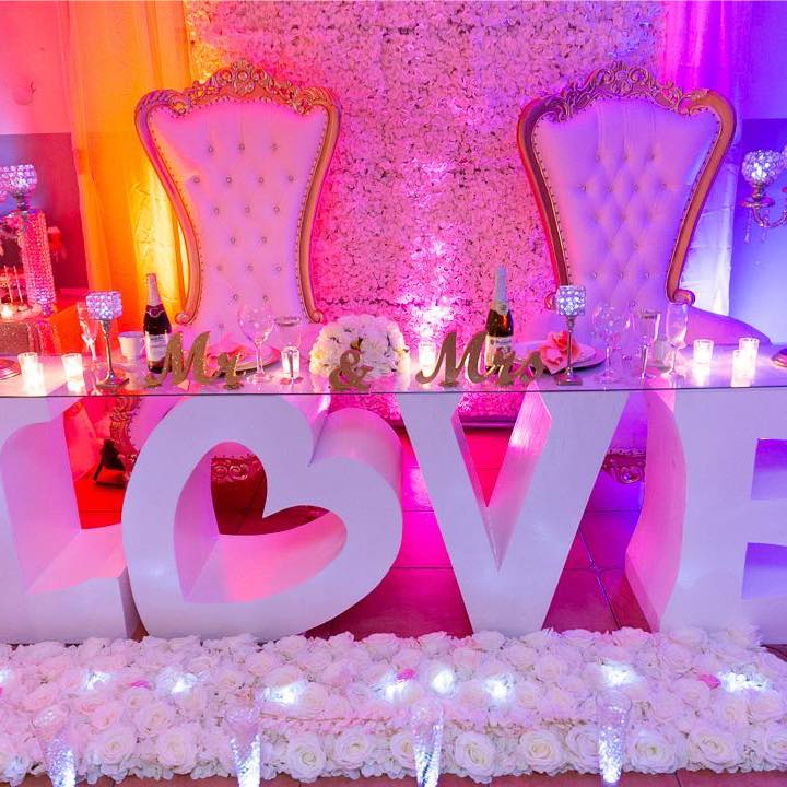 Wedding sweetheart table with white floral wall, gold thrones, and large LOVE letters with rose accents